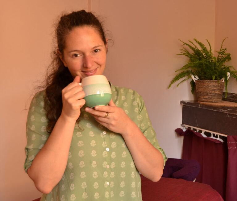 Praticienne en ayurveda assise sur la table de massage, elle regarde la caméra avec une tasse de tisane dans les mains et un sourire