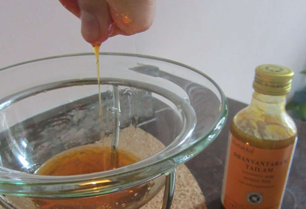 Main qui fait couler un filet d'huile dans un chauffe-huile chauffé grâce à une bougie. Une bouteille d'huile Dhanwantaram est à côté